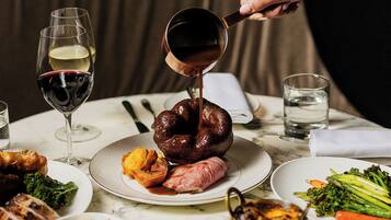 A dining table with plates of food, a glass of red wine, and a person pouring a dark liquid into a bowl.