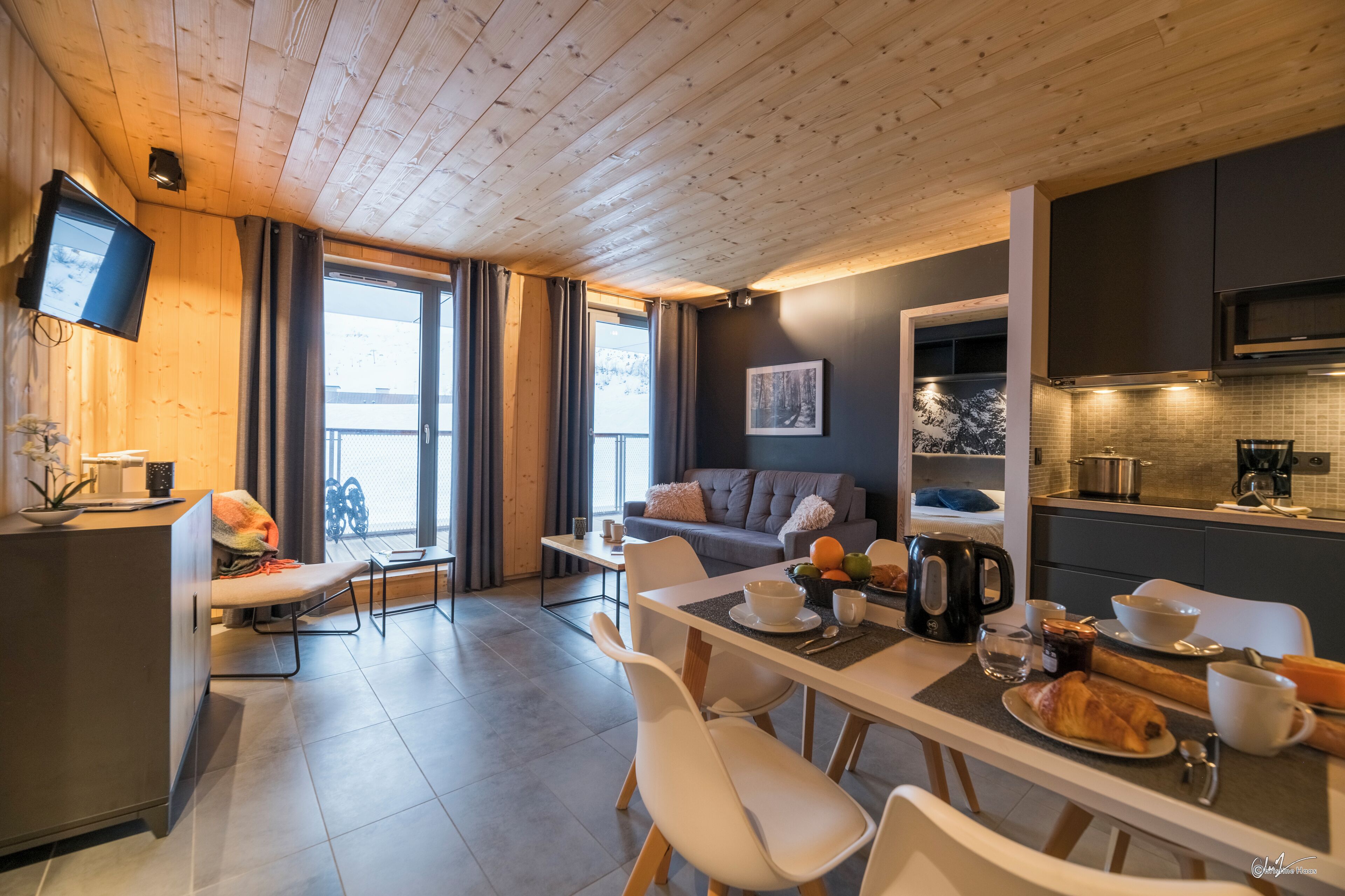 A modern kitchen with a dining area, a sofa, and a TV mounted on the wall.