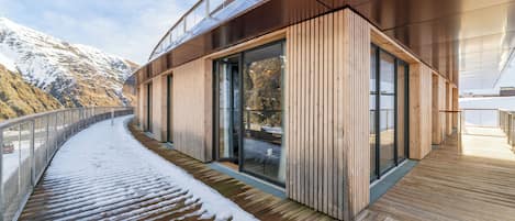 A modern building with a wooden deck and glass doors, overlooking a snowy mountain.