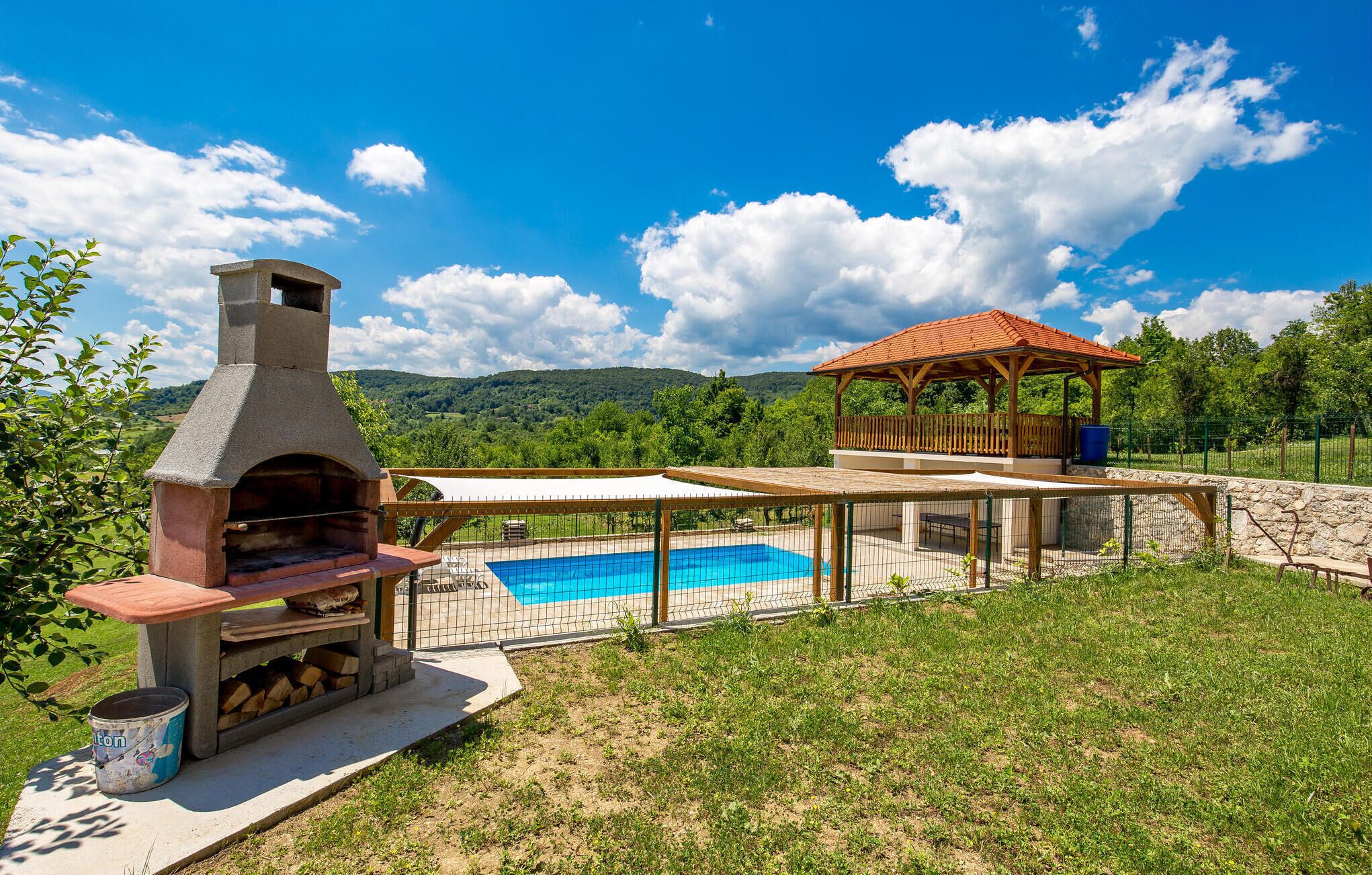 A backyard with a barbecue grill, a swimming pool, a wooden gazebo, and a stone wall.