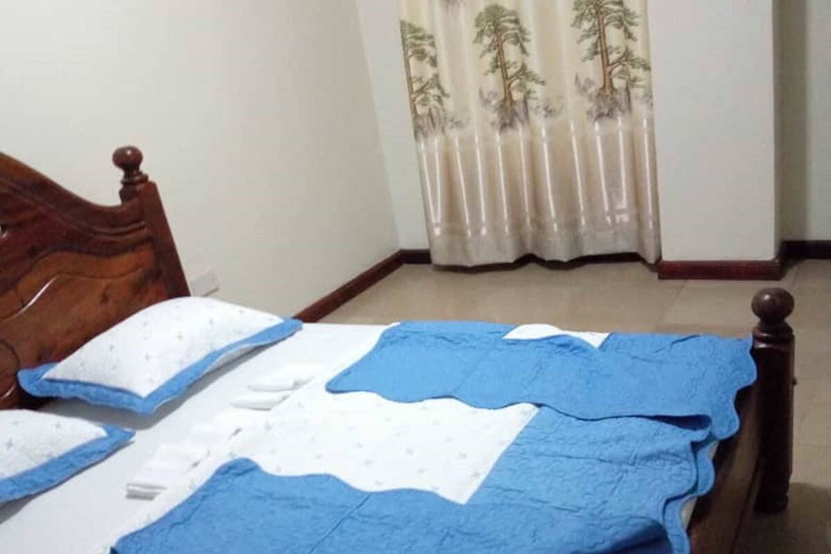 Un dormitorio con estructura de cama de madera, colcha azul y blanca, y una ventana con cortinas ligeras.