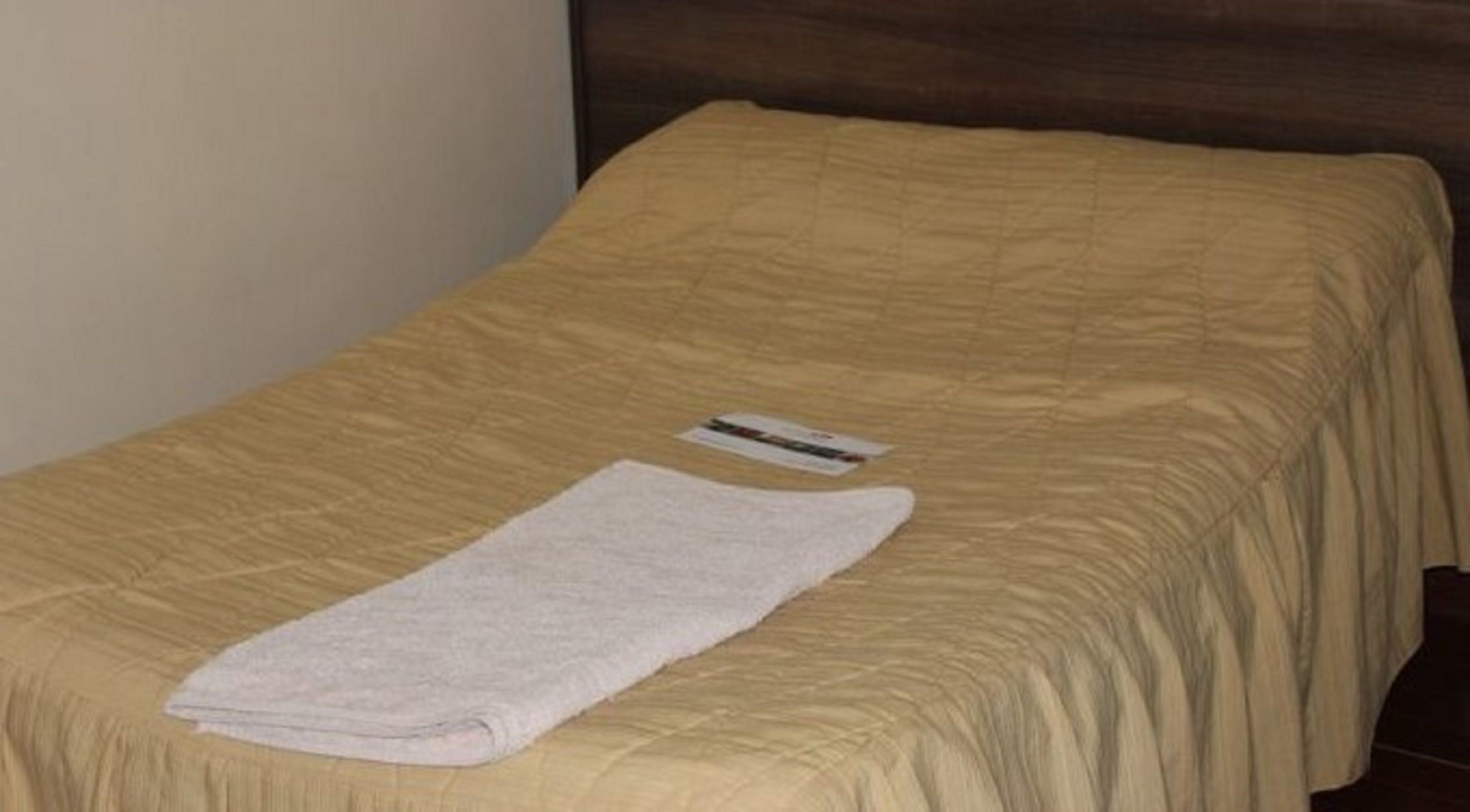 Una cama individual con una colcha color crema y una toalla blanca doblada encima.