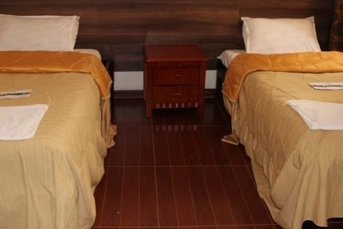 Een kamer met twee bedden, elk met een wit kussen en een beige deken. Daartussen bevindt zich een houten nachtkastje met een lade.