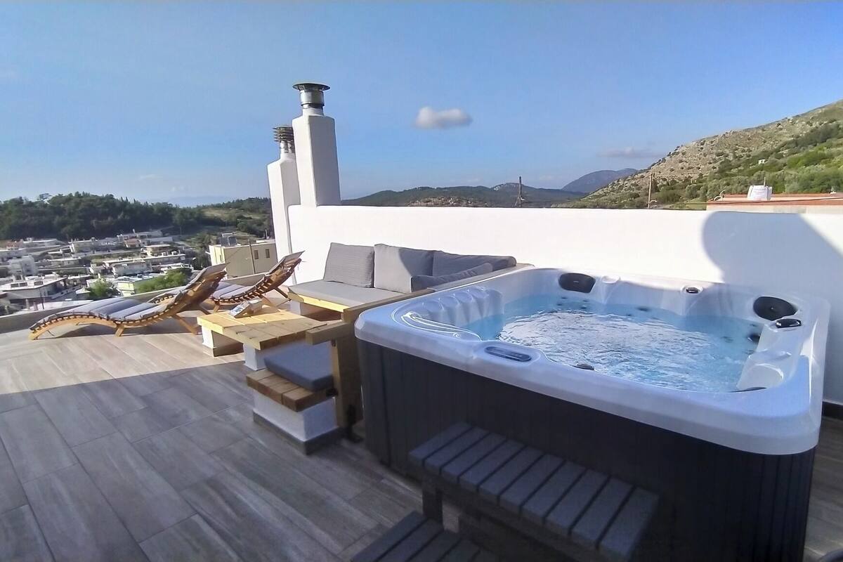 A rooftop terrace with a hot tub, lounge chairs, and a view of a town and hills.