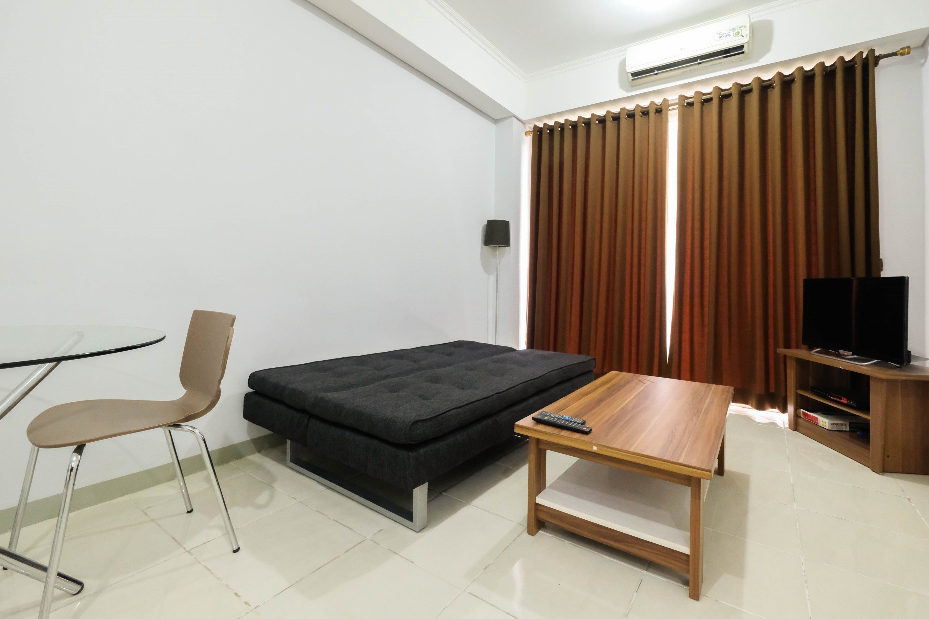 A compact living space with a sofa bed, a small wooden table, a chair, a TV stand, and a window with curtains.