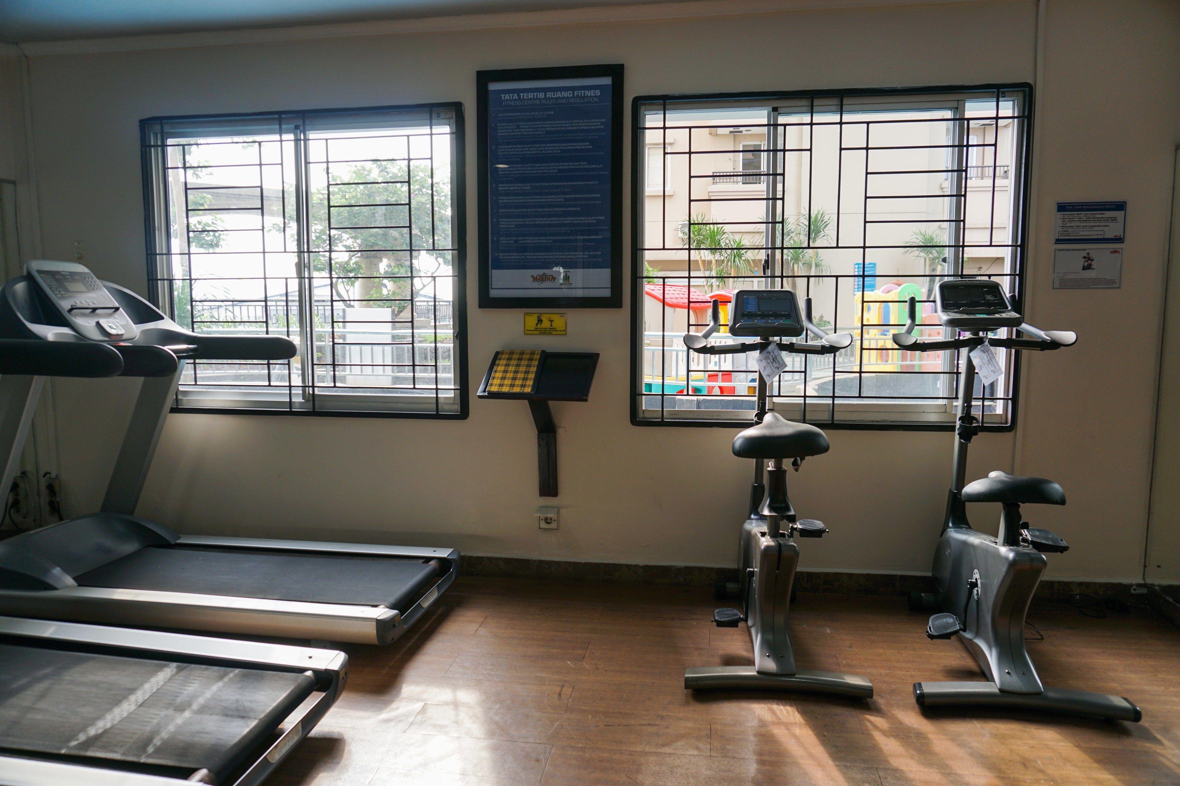 Un gimnasio con cintas de correr y bicicletas estáticas, ventanas amplias con barrotes de metal y un cartel en la pared.