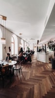 A spacious restaurant with high ceilings and wooden flooring, featuring tables and chairs arranged for dining.