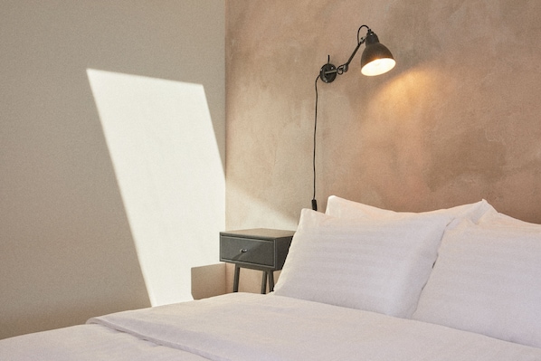 Een net opgemaakt bed met witte lakens, een nachtkastje met een lamp, en een wand met een structuur.