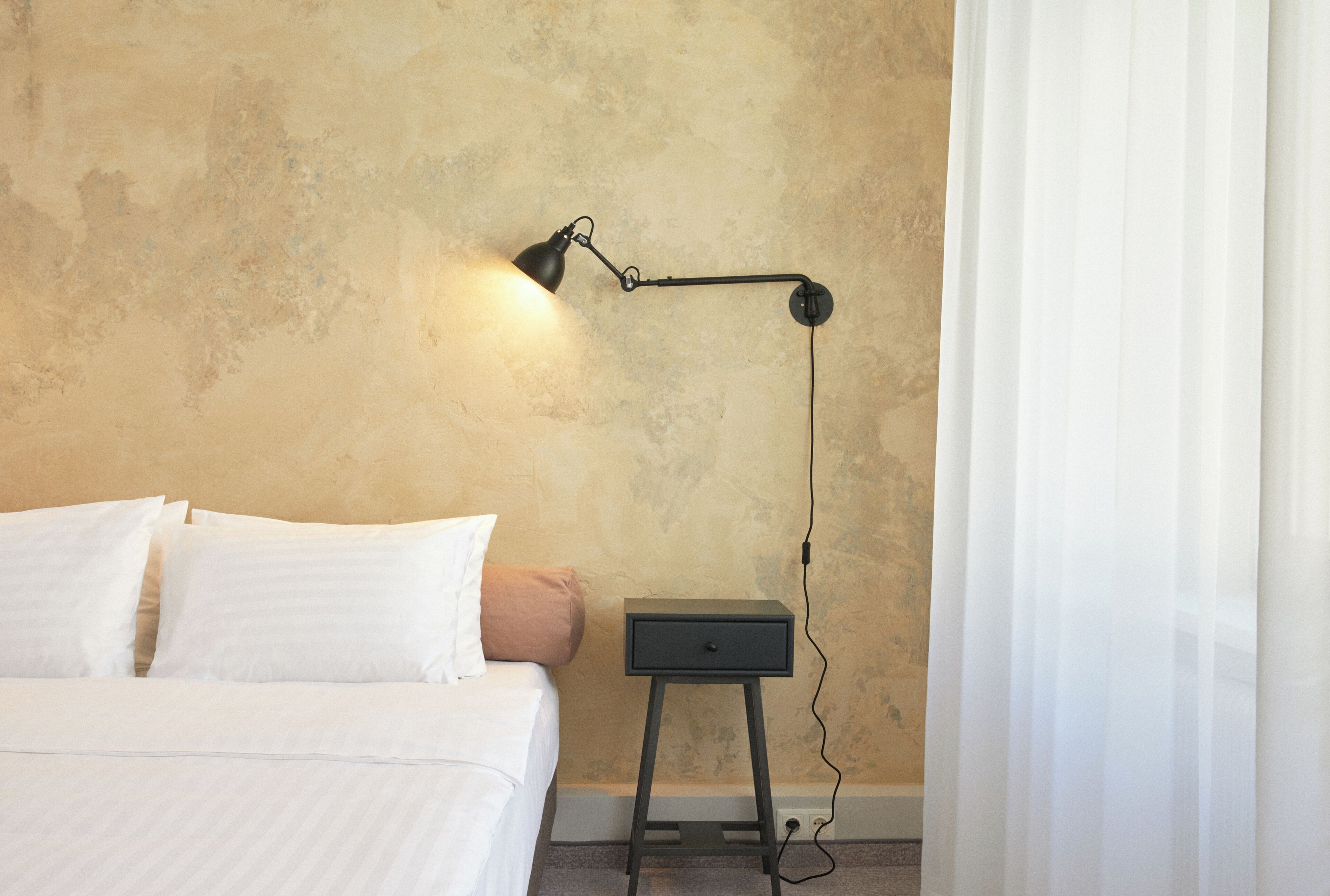 Een slaapkamer met een bed, kussens, een nachtkastje, een lamp en een stopcontact.