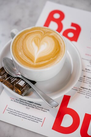 A cup of coffee with latte art on a saucer, a spoon, and a menu with red text. - Hotel Babula am Augarten (Vienna)
