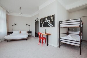 A hotel room with a bed, a desk with a chair, a bunk bed, and a painting on the wall.