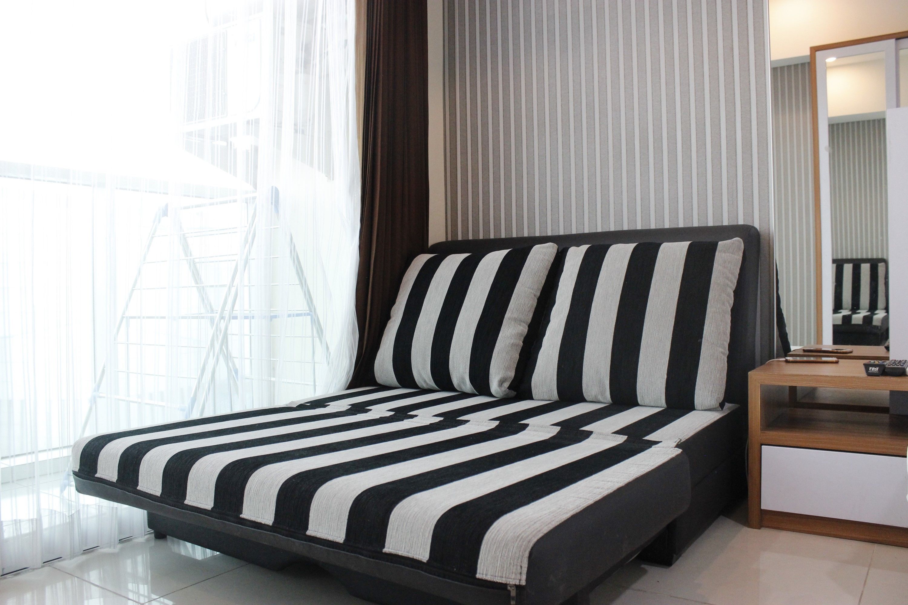Ein modernes Schlafzimmer mit einem schwarz-weiß gestreiften Bett, einem Holfnachtkästchen und einem großen Fenster mit durchsichtigen Vorhängen.