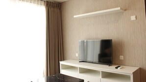 A modern living room with a flat-screen TV on a white stand, beige curtains, and an air conditioning unit on the wall. - Classic 2BR Apartment At Gateway Pasteur near Exit Toll (Cimahi)
