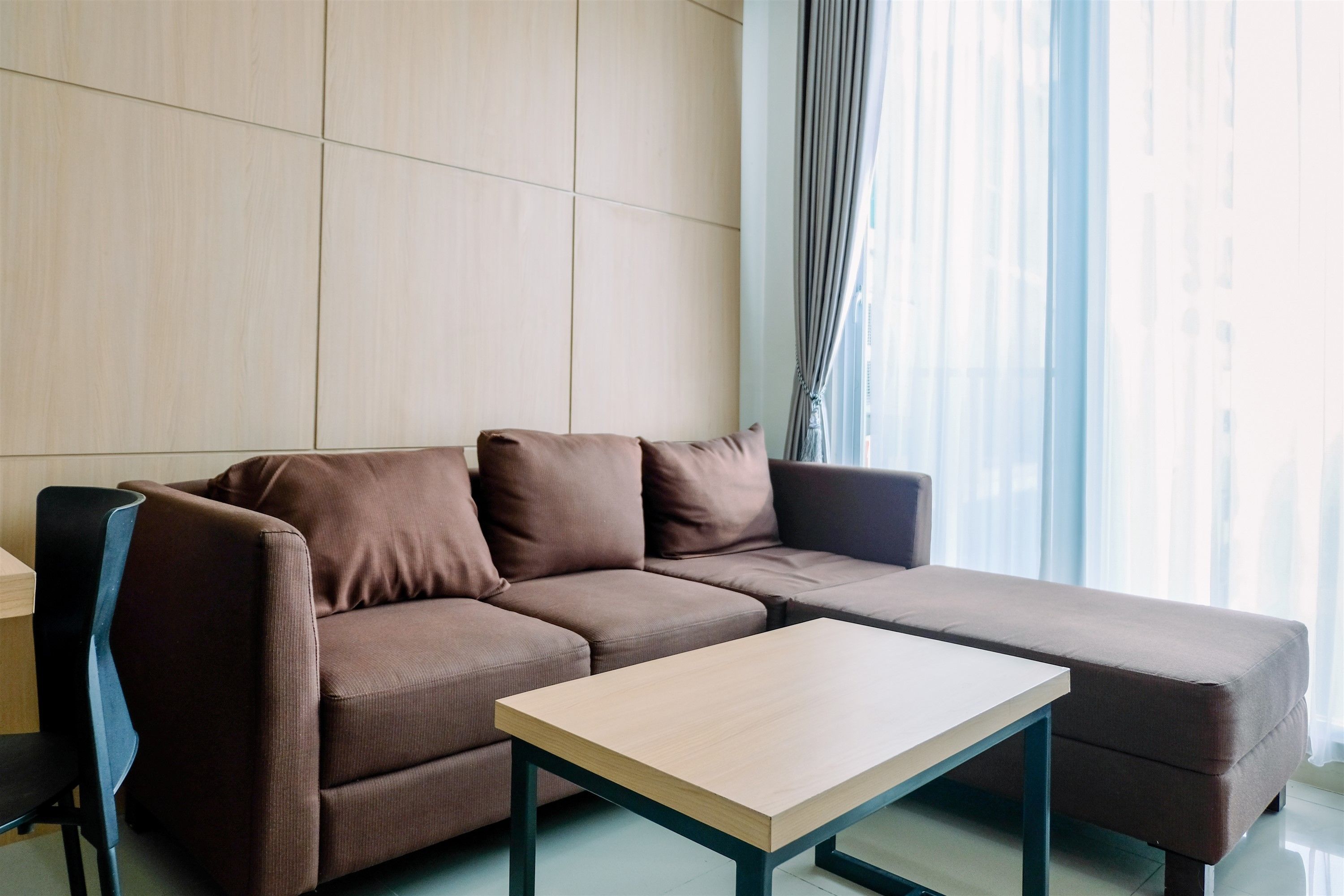 A modern living room with a brown sectional sofa, a wooden coffee table, and a view of a cityscape through sheer curtains.