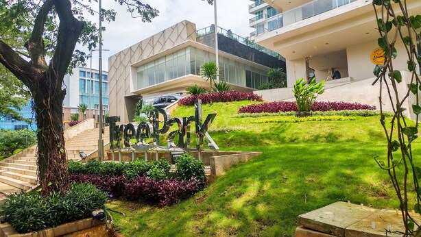 A modern building with a glass facade, a sign reading "TODAY," and a well-maintained garden with various plants and a tree. - Comfortable 1BR Tree Park Apartment (South Tangerang)