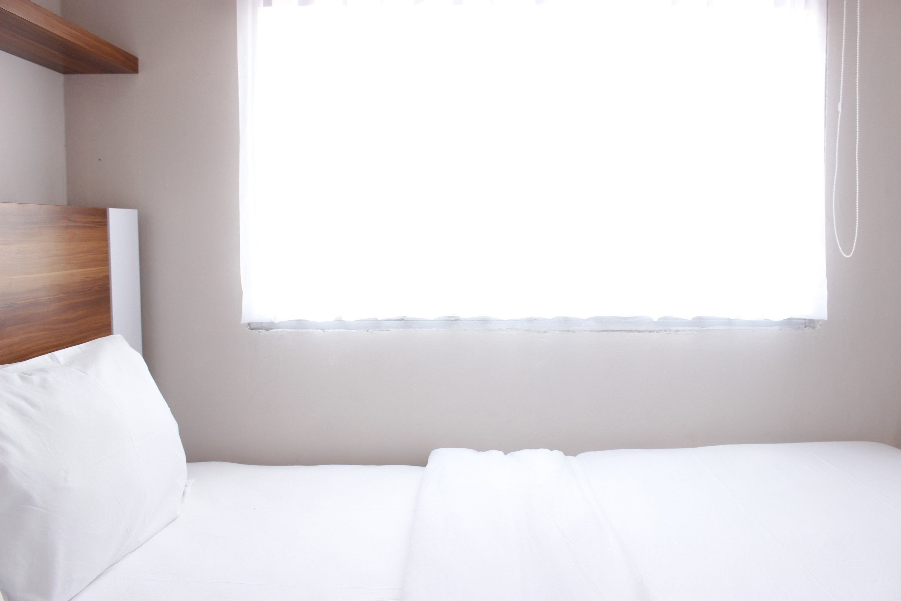 Un lit simple avec une tête de lit en bois, un oreiller blanc et une fenêtre ornée d’un rideau léger.