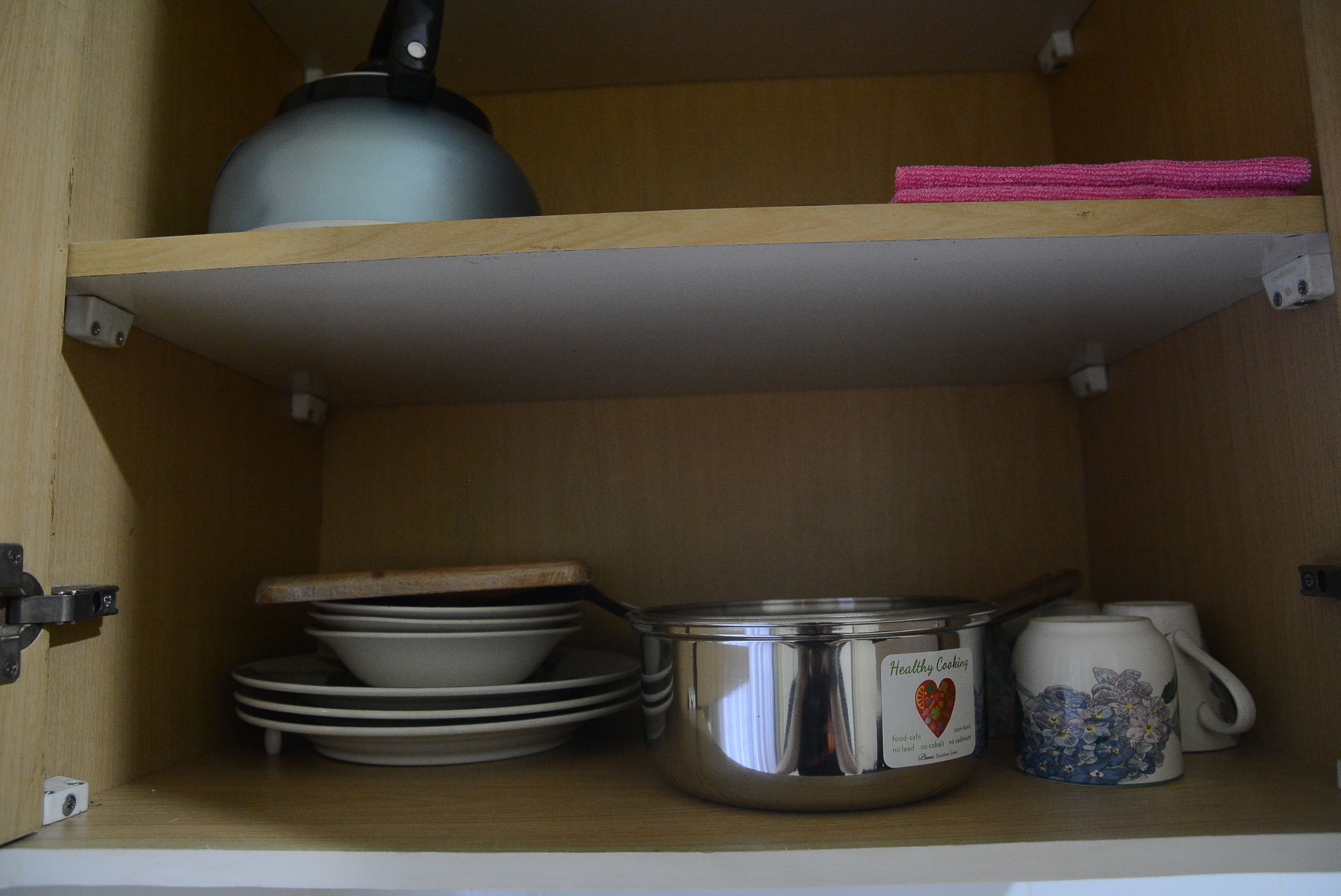 A kitchen cabinet with a kettle, pots, plates, and a mug.