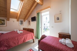 A hotel room with two beds, wooden ceiling beams, a TV, and a view of a church through a window. - R&B La Gerla Blu (Verzegnis)