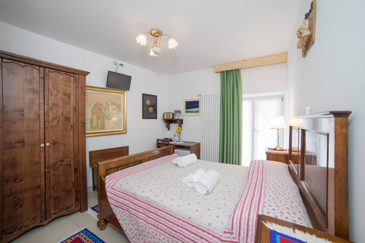 A bedroom with a wooden wardrobe, a bed with a pink and white striped bedspread, a television on the wall, and a green curtain.