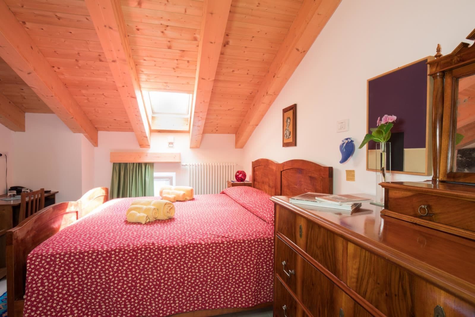 Een slaapkamer met een houten plafond, een bed met een bloemenprint beddeken, een dressoir en een spiegel.
