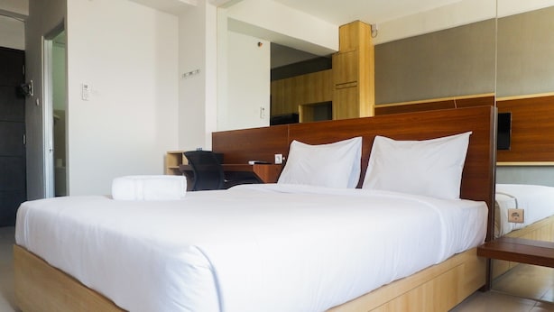 A modern hotel room with a large bed, wooden headboard, and a small desk with a chair. - Comfy Studio Apartment at Menara Rungkut (Surabaya)