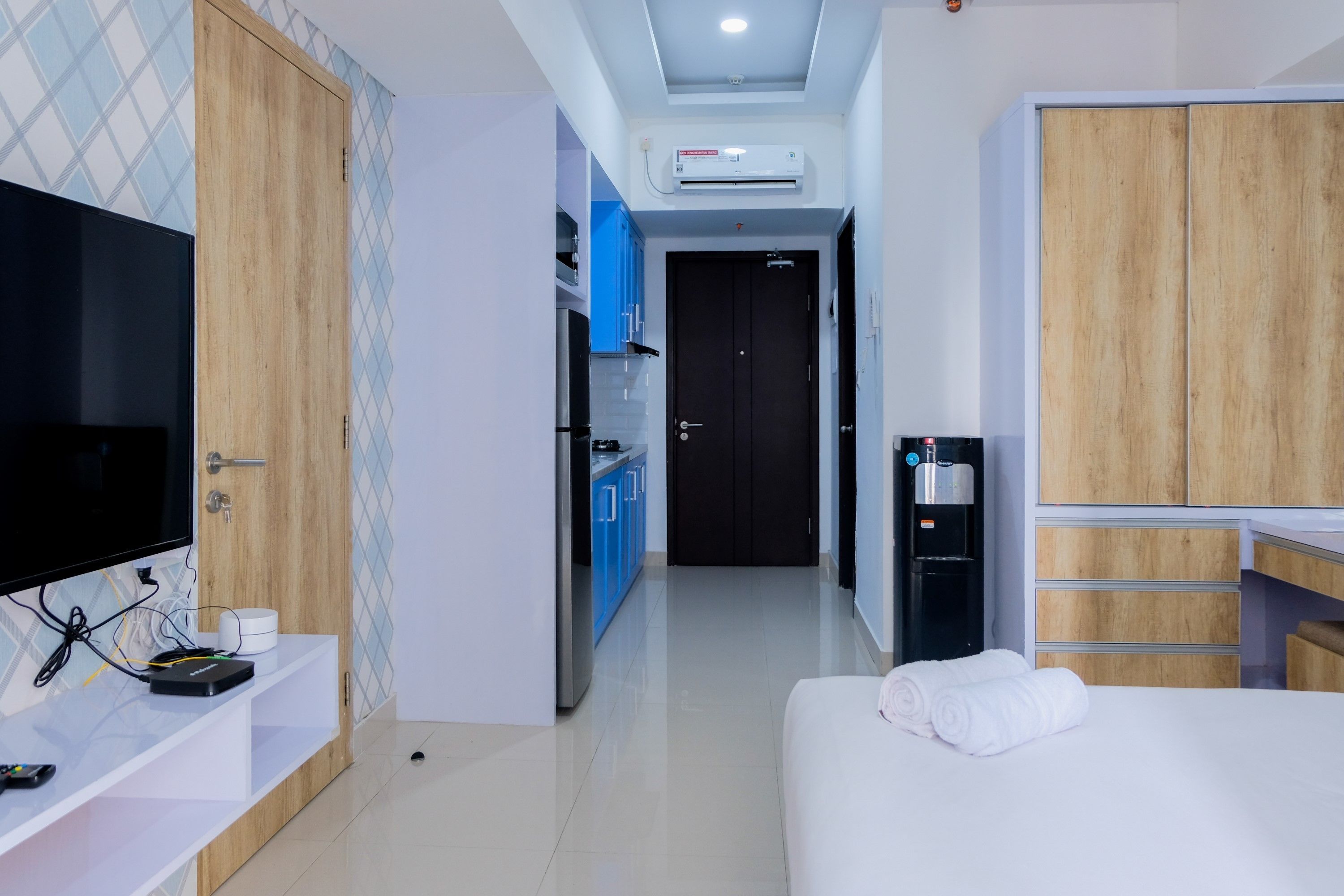 A hotel room with a bed, a TV, a desk, a wardrobe, a kitchenette, and an air conditioning unit.