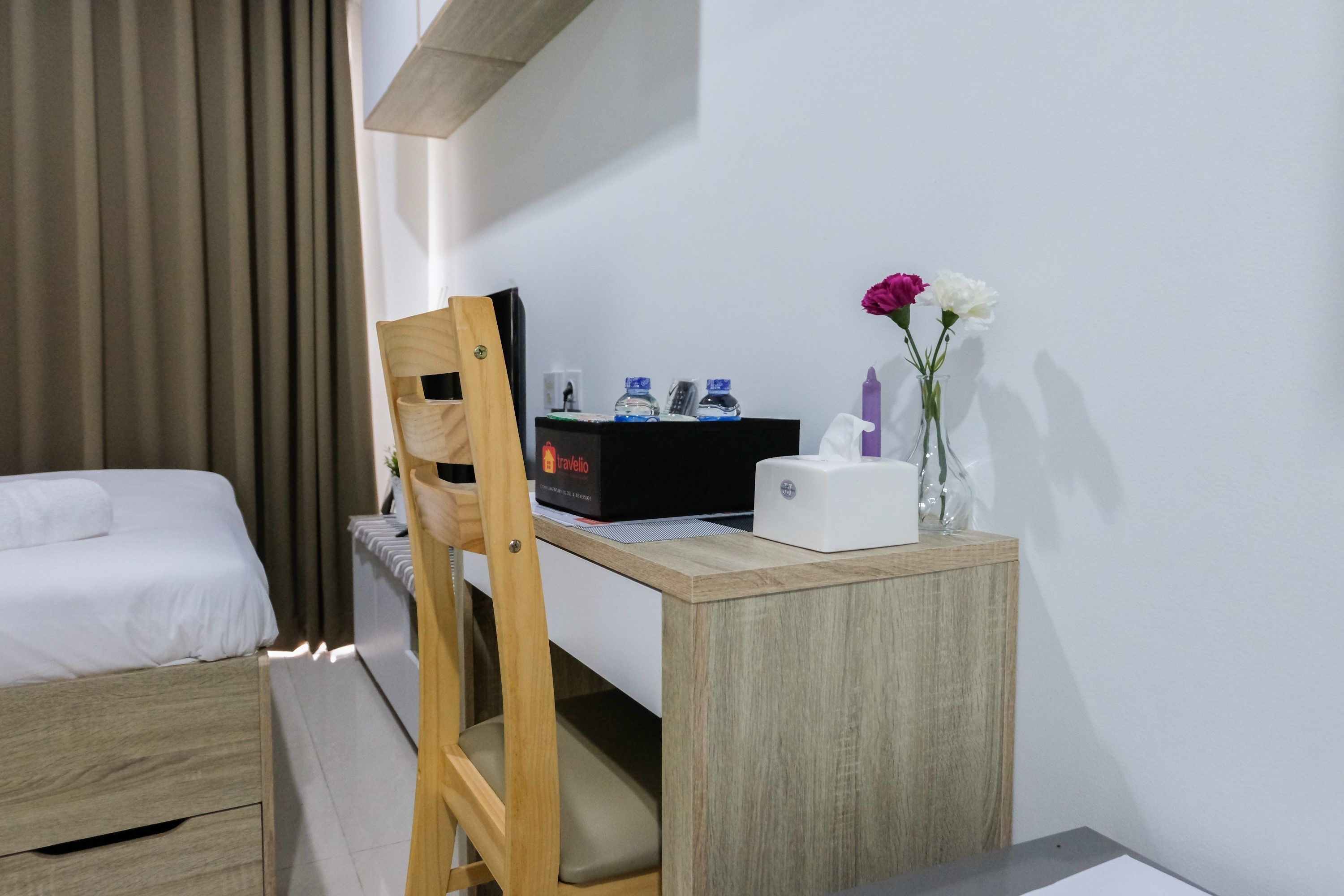 A hotel room with a bed, a desk with a computer, a chair, a small table with a vase of flowers, and a wall-mounted telephone.