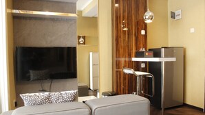 Une chambre d’hôtel moderne dotée d’un téléviseur à écran plat fixé au mur, d’une kitchenette avec réfrigérateur et d’un coin salon comprenant un canapé et une table basse.