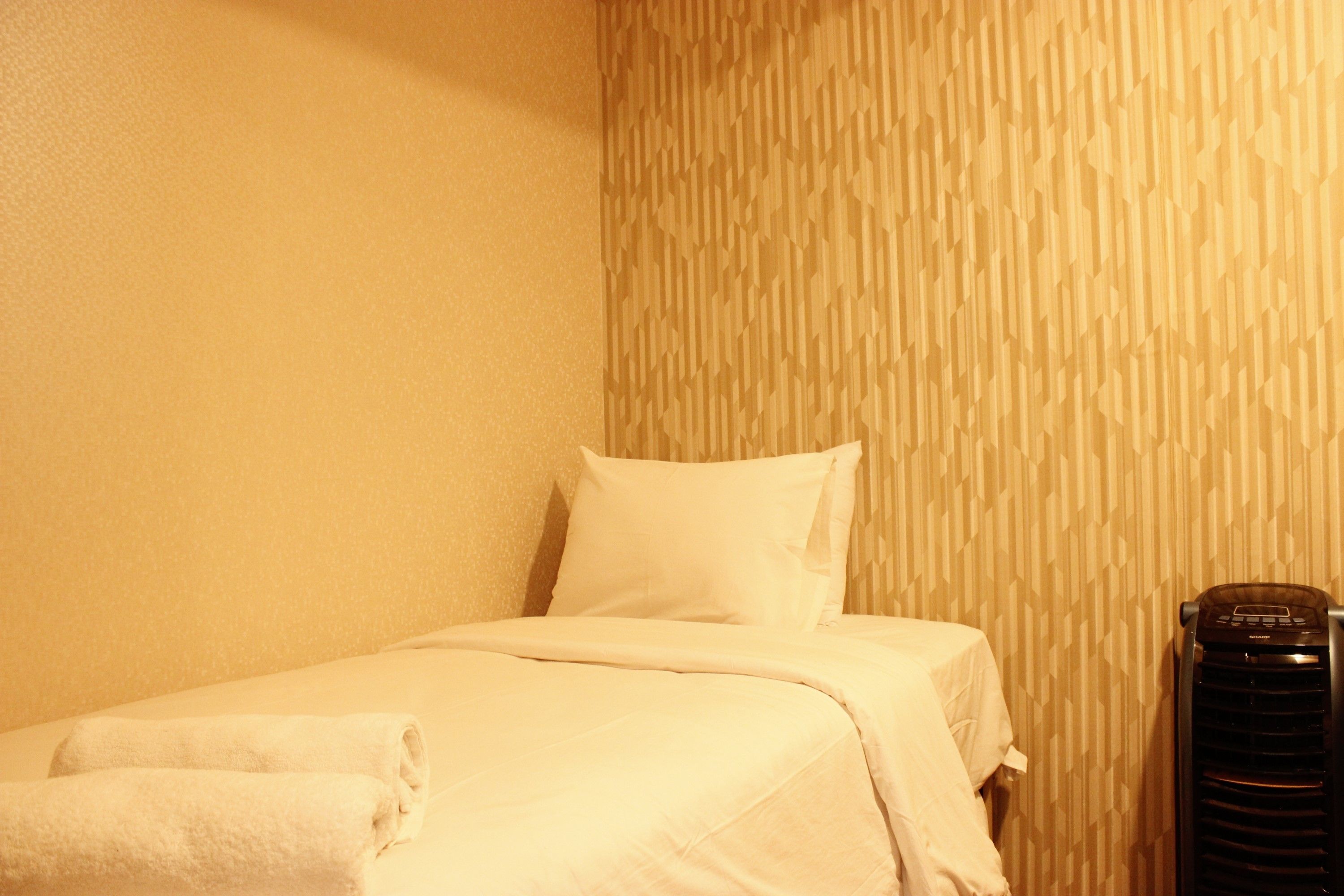 A neatly made bed with white linens and a rolled towel, a black heater, and a textured wall pattern.