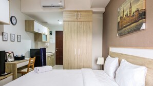 A hotel room with a bed, a desk with a chair, a wardrobe, a painting on the wall, and an air conditioning unit.
