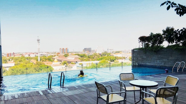 Una piscina exterior con terraza de madera, sillas de descanso y vistas a un paisaje urbano de fondo.