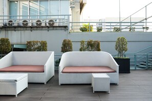 A rooftop terrace with two white wicker sofas with brown cushions, a small table, and potted plants.