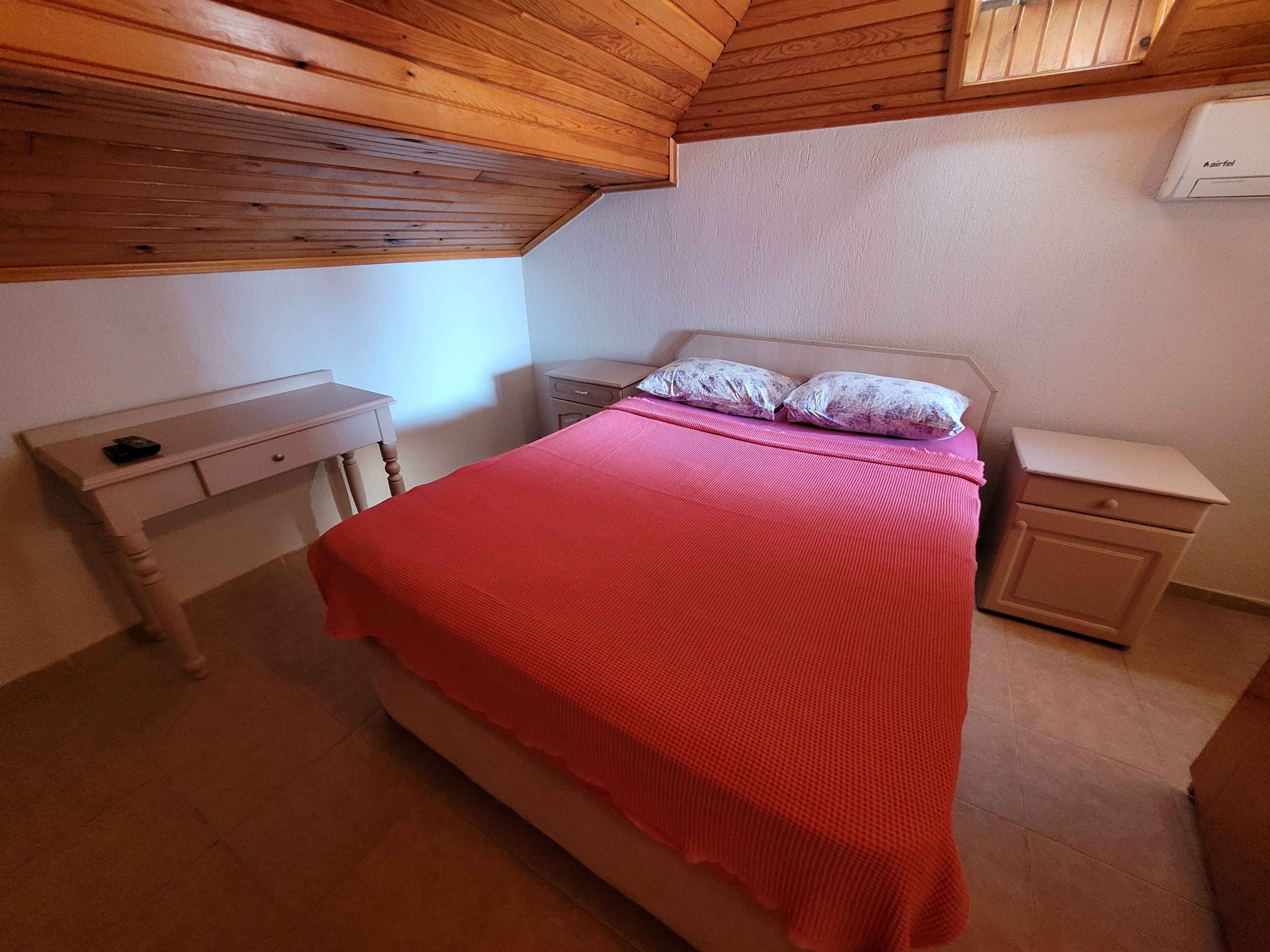 A bedroom with a wooden ceiling, a bed with a red blanket, two bedside tables, and a nightstand.