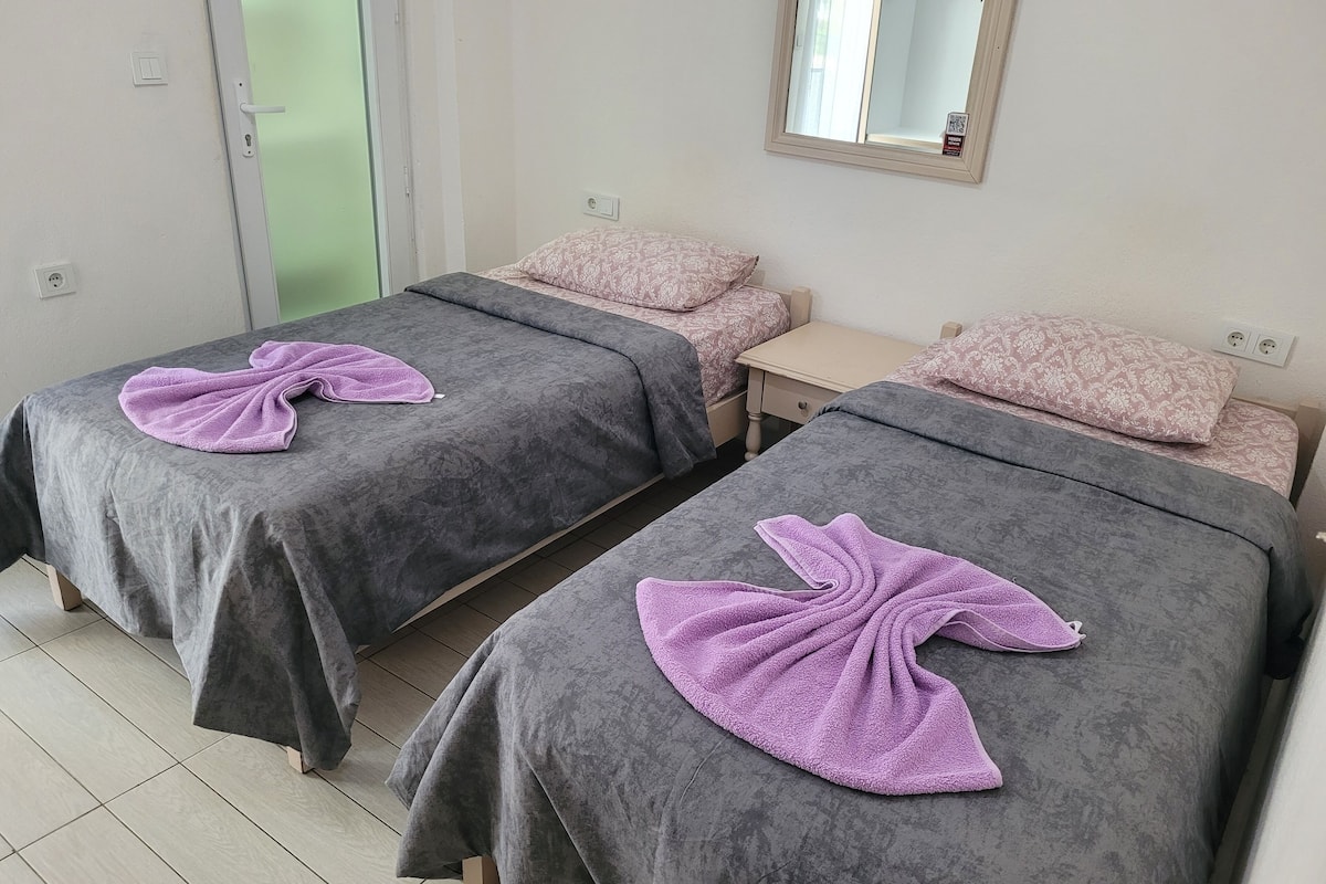 Two twin beds with grey linens and purple towels, a small bedside table, a mirror, and a green door.