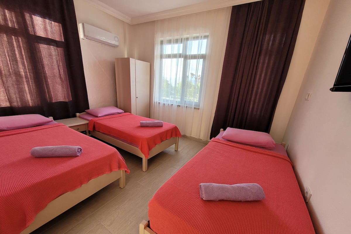 A hotel room with three single beds, each with a red bedspread and a rolled towel. There is a wardrobe, a window with curtains, and an air conditioning unit on the wall.