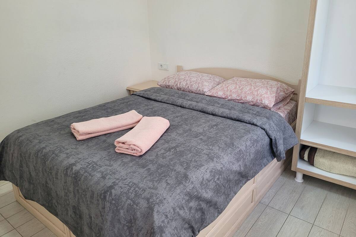 A neatly made bed with a grey bedspread and two pink towels, a wooden nightstand with a lamp, and a white wall.