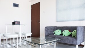 Un soggiorno moderno con un tavolino in vetro, sedie bianche, un divano grigio con peluche verdi e una porta marrone.