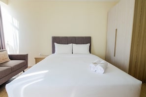 A neatly made bed with white linens and two pillows, a wooden wardrobe to the right, and a small bedside table with a lamp.