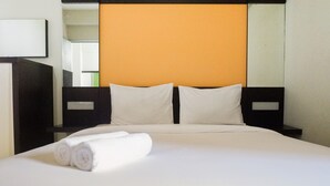 A neatly made bed with white linens and rolled towels, a black headboard, and a plain orange wall behind it.