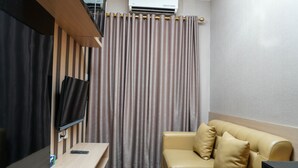 Una habitación de hotel moderna con televisor de pantalla plana, un escritorio de madera, un sofá color beige y una cortina grande.