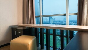 Una habitación de hotel moderna con un ventanal grande que ofrece vistas a la ciudad, un escritorio pequeño y una zona de estar con cojines.