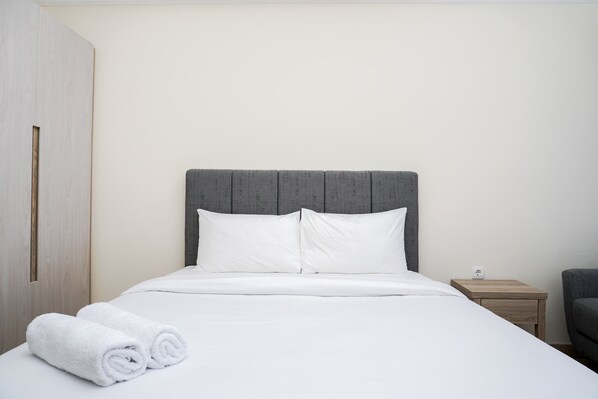 A neatly made bed with white linens and pillows, a wooden nightstand with a lamp, and a grey upholstered chair.