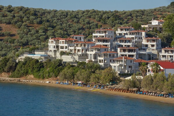 A coastal residential area with multi-story buildings, a beach with sun loungers, and a clear view of the sea. - Olivia Sports and Sea (Almyros)