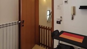 Una habitación con puerta de madera, un radiador, una mesa con dos sillas y un estante pequeño con objetos decorativos.