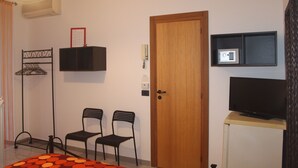 Una habitación con puerta de madera, televisor sobre un pedestal, dos sillas, un estante con una caja fuerte y una lámpara colgante.