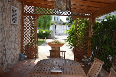 A wooden pergola with a table and chairs, surrounded by potted plants and a stone wall.