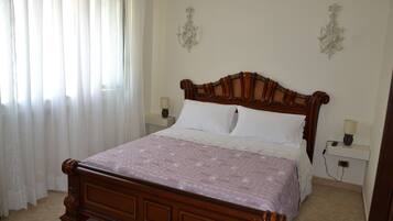 Un letto rifatto con cura, con una testiera in legno, cuscini bianchi e una coperta a motivi. Sono presenti due comodini con lampade, e la stanza ha un pavimento piastrellato.
