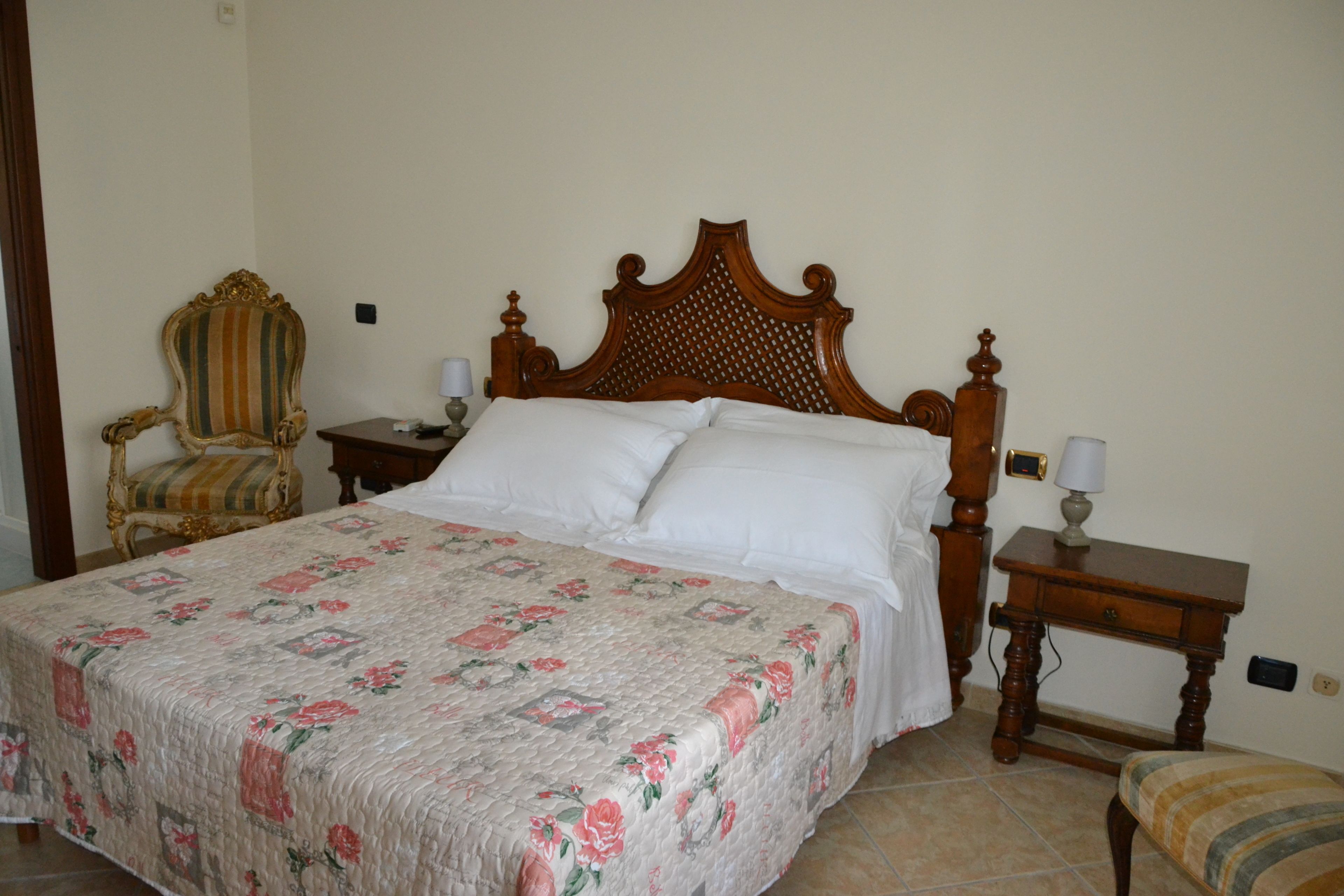 A neatly made bed with a floral bedspread, a wooden headboard, a bedside table with a lamp, a chair, and a small stool.