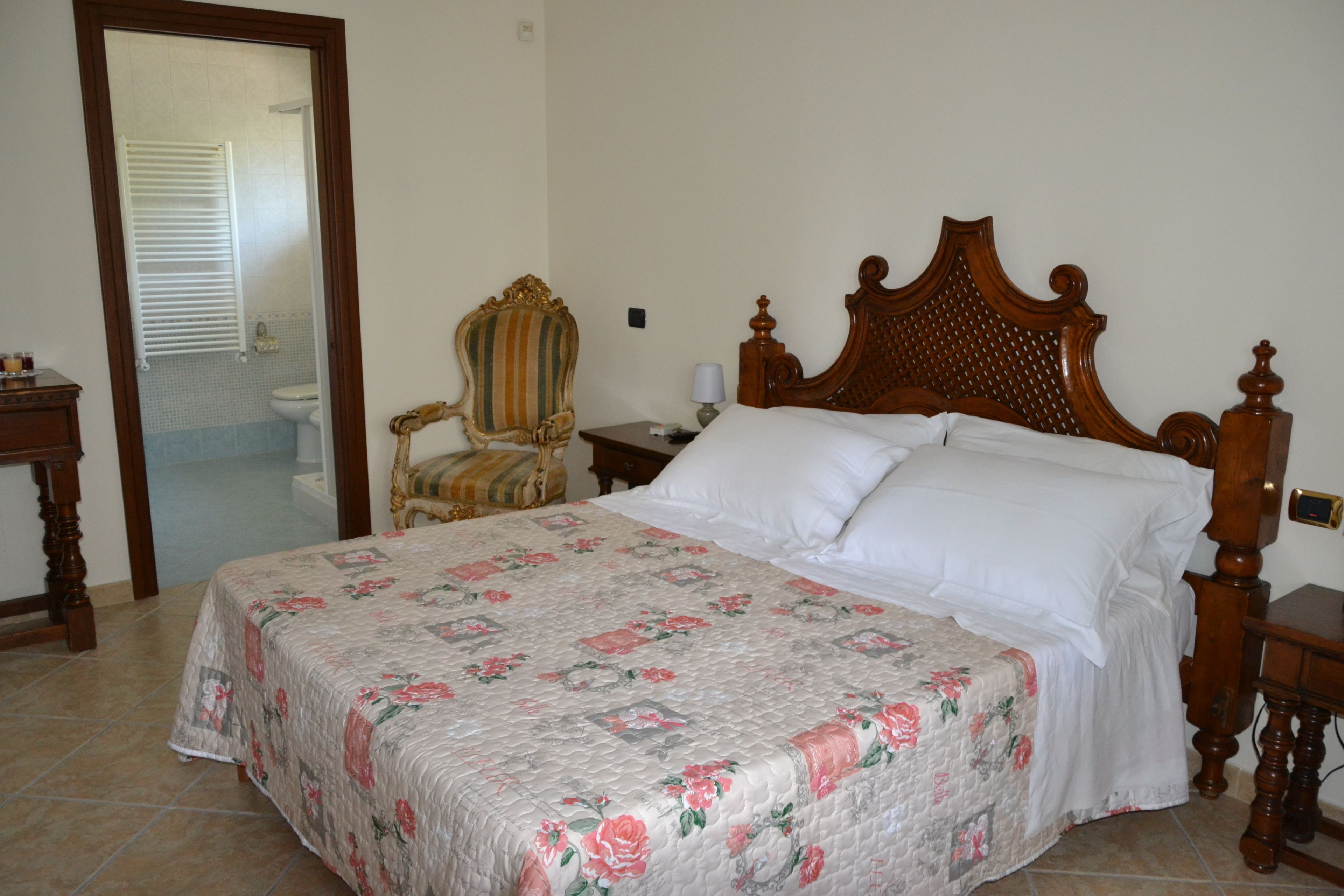 Ein Schlafzimmer mit einem hölzernen Bett, einem Sessel, einem Nachttisch und einem durch eine geöffnete Tür einsehbarer Badezimmer.