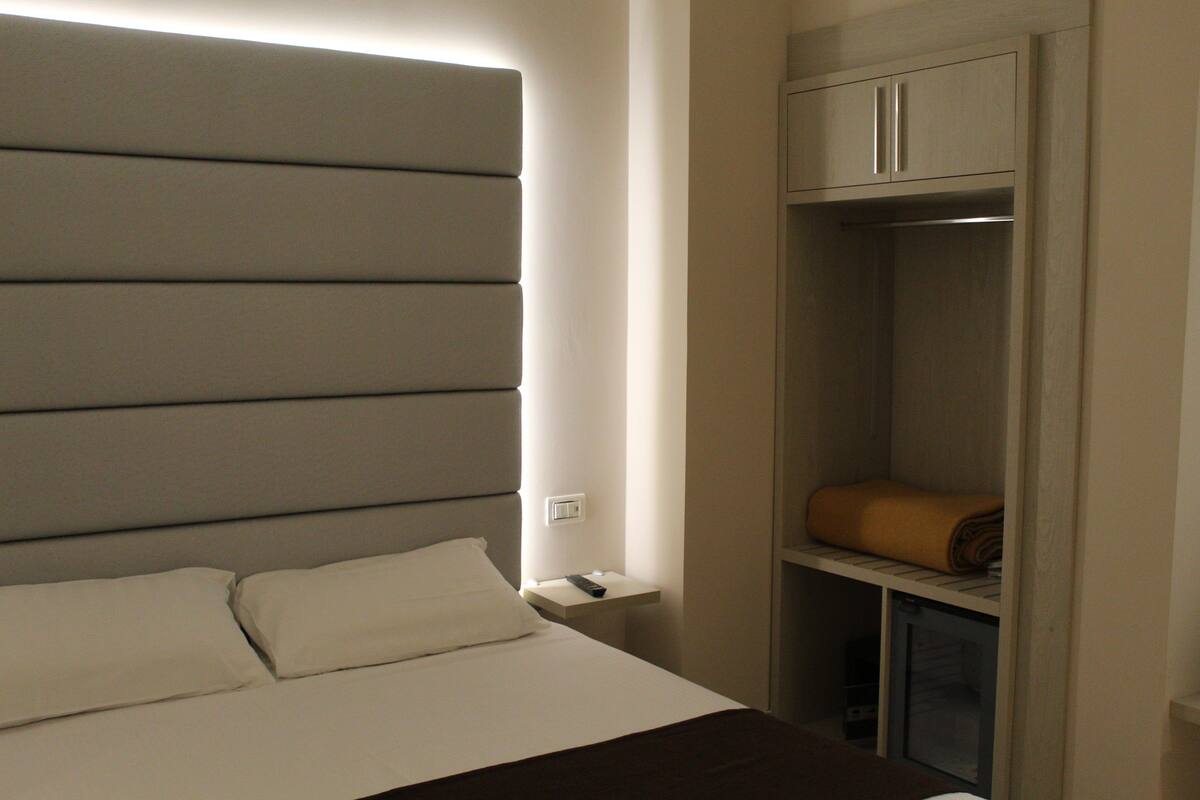 Ein Hotelzimmer mit Bett, Nachttisch, Kleiderschrank und Ablagefläche mit zusammengerolltem Handtuch.